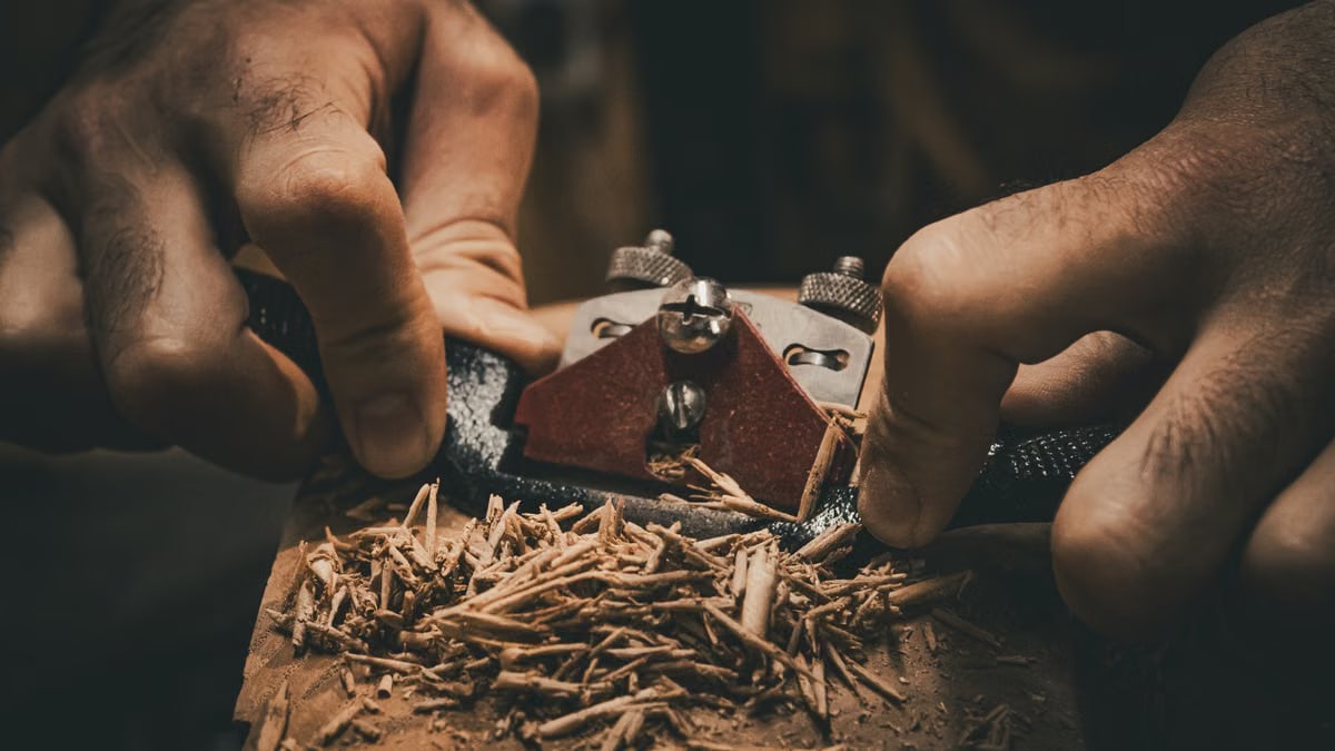 Detailed view of hands using a hand plane tool in woodworking, creating wood shavings.